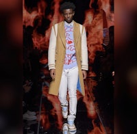 a man walks down the runway in a tan coat and white pants
